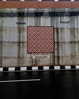 The image features a concrete wall with a decorative batik pattern in the center, set above a roadway with a black and white striped curb. There is text beneath the pattern, reading 'BATIK CEPLOK SEKAR KOPI.' Water stains are visible on the concrete surface, indicating possible weather exposure.