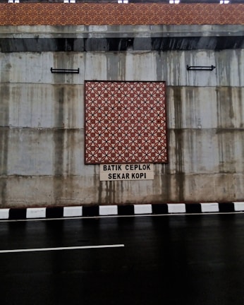 The image features a concrete wall with a decorative batik pattern in the center, set above a roadway with a black and white striped curb. There is text beneath the pattern, reading 'BATIK CEPLOK SEKAR KOPI.' Water stains are visible on the concrete surface, indicating possible weather exposure.