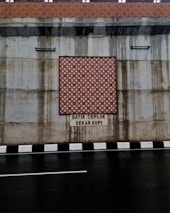 The image features a concrete wall with a decorative batik pattern in the center, set above a roadway with a black and white striped curb. There is text beneath the pattern, reading 'BATIK CEPLOK SEKAR KOPI.' Water stains are visible on the concrete surface, indicating possible weather exposure.