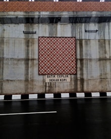 The image features a concrete wall with a decorative batik pattern in the center, set above a roadway with a black and white striped curb. There is text beneath the pattern, reading 'BATIK CEPLOK SEKAR KOPI.' Water stains are visible on the concrete surface, indicating possible weather exposure.