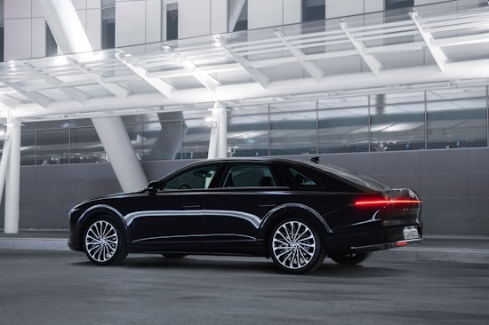 A sleek black luxury sedan is parked under a modern, well-lit structure with large glass panels and white beams. The car&rsquo;s design features aerodynamic lines and prominently lit rear lights.