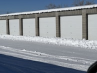 A row of storage unit garages with closed white doors, surrounded by snow on the ground and roof. The sky is clear and blue, indicating a cold winter day.