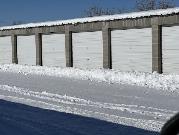 A row of storage unit garages with closed white doors, surrounded by snow on the ground and roof. The sky is clear and blue, indicating a cold winter day.