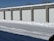 A row of storage unit garages with closed white doors, surrounded by snow on the ground and roof. The sky is clear and blue, indicating a cold winter day.