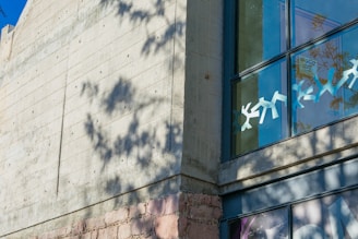 A modern concrete building facade with a large window displaying geometric paper cutout decorations. The shadow of a nearby tree creates an intricate pattern on the wall. Part of a faded mural or artwork is visible at the bottom right corner.