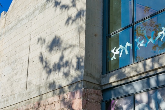 A modern concrete building facade with a large window displaying geometric paper cutout decorations. The shadow of a nearby tree creates an intricate pattern on the wall. Part of a faded mural or artwork is visible at the bottom right corner.