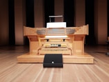 A large wooden pipe organ console is situated in a performance hall, featuring multiple rows of keys and a variety of knobs and pedals. The organ is positioned on a wooden stage with a backdrop of tall, rectangular wooden panels.