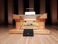 A large wooden pipe organ console is situated in a performance hall, featuring multiple rows of keys and a variety of knobs and pedals. The organ is positioned on a wooden stage with a backdrop of tall, rectangular wooden panels.