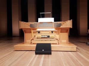 A large wooden pipe organ console is situated in a performance hall, featuring multiple rows of keys and a variety of knobs and pedals. The organ is positioned on a wooden stage with a backdrop of tall, rectangular wooden panels.