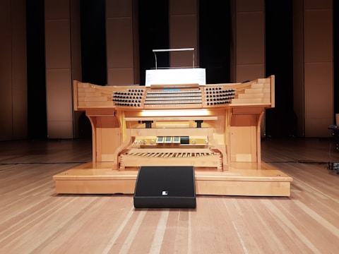A large wooden pipe organ console is situated in a performance hall, featuring multiple rows of keys and a variety of knobs and pedals. The organ is positioned on a wooden stage with a backdrop of tall, rectangular wooden panels.