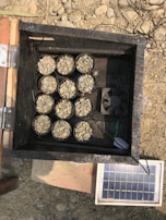 A wooden box with open top contains nine small containers filled with pebbles. A small fan is also visible inside the box. Outside the box, there is a small solar panel connected by wires, resting on rocky ground.