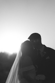 A couple is silhouetted against a bright light, likely the setting sun. The bride wears a veil and holds a bouquet, while the groom is dressed in a suit. Their figures are dark against the bright background, creating a romantic and timeless atmosphere.