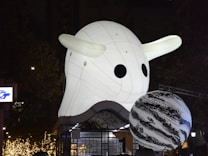A large, illuminated inflatable sculpture resembling a cartoonish ghost with dark round eyes is prominently displayed in an urban nighttime setting. Nearby, a spherical object with swirling black and white patterns adds to the playful scene. Twinkling lights and dark foliage can be seen in the background.