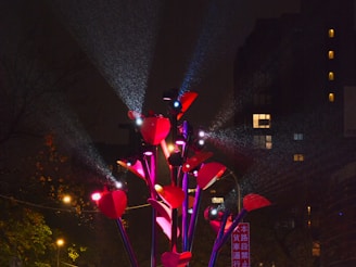 Brightly colored red and pink light installation resembling flowers or leaves is prominently displayed against a dark urban background. Multiple beams of light are directed from the installation towards the sky, creating a vibrant visual effect. Surrounding buildings and trees are dimly lit with scattered illuminated windows, adding to the atmospheric setting.
