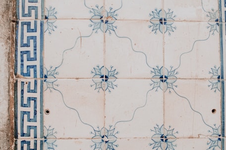 Ornate ceramic tiles featuring a floral pattern with blue designs. The tiles display a symmetrical design with a border on the left side showcasing a geometric motif. The overall look is rustic with slight imperfections visible.
