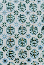 A pattern of ceramic tiles consists of circular designs with crisscrossing green lines against a cream background. Each circle encloses a small architectural shape resembling a building, and the circles are framed by star-like shapes with alternating blue and green petals.