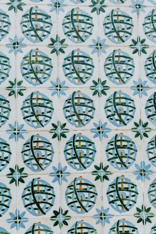 A pattern of ceramic tiles consists of circular designs with crisscrossing green lines against a cream background. Each circle encloses a small architectural shape resembling a building, and the circles are framed by star-like shapes with alternating blue and green petals.