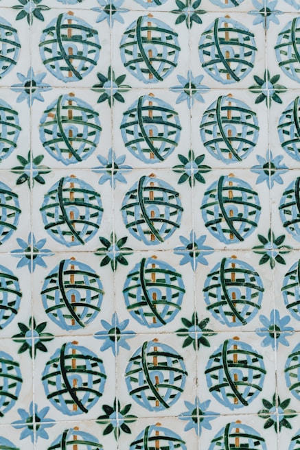 A pattern of ceramic tiles consists of circular designs with crisscrossing green lines against a cream background. Each circle encloses a small architectural shape resembling a building, and the circles are framed by star-like shapes with alternating blue and green petals.