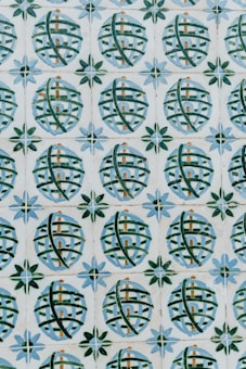 A pattern of ceramic tiles consists of circular designs with crisscrossing green lines against a cream background. Each circle encloses a small architectural shape resembling a building, and the circles are framed by star-like shapes with alternating blue and green petals.