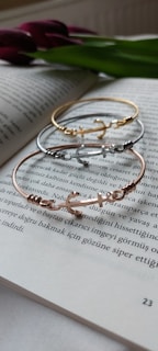 A trio of delicate bracelets with an anchor design lay atop an open book. The book’s text is in Turkish and is in focus, adding an intellectual ambiance. In the background, a green leaf and purple flowers are slightly blurred, giving a touch of nature.