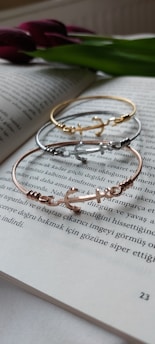 A trio of delicate bracelets with an anchor design lay atop an open book. The book’s text is in Turkish and is in focus, adding an intellectual ambiance. In the background, a green leaf and purple flowers are slightly blurred, giving a touch of nature.