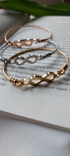 Three metal bracelets with infinity symbols are laid on the pages of an open book. The bracelets are in gold, silver, and rose gold tones, positioned in a slightly overlapping manner. The background includes blurred text in a foreign language.