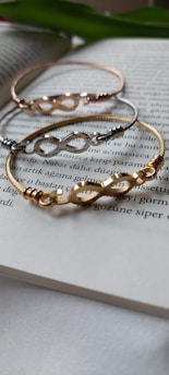 Three metal bracelets with infinity symbols are laid on the pages of an open book. The bracelets are in gold, silver, and rose gold tones, positioned in a slightly overlapping manner. The background includes blurred text in a foreign language.