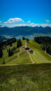 A scenic view of a lush green valley with a narrow dirt path leading to a small farmhouse surrounded by trees. In the background, there are rolling hills and a distant lake, with mountains and a clear blue sky featuring a few fluffy clouds.