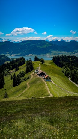 A scenic view of a lush green valley with a narrow dirt path leading to a small farmhouse surrounded by trees. In the background, there are rolling hills and a distant lake, with mountains and a clear blue sky featuring a few fluffy clouds.