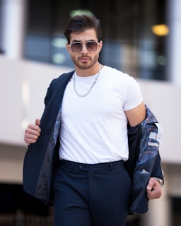 A man in sunglasses and a white t-shirt poses confidently while holding a suit jacket over his shoulder. He wears a chain necklace and dark pants, exuding a stylish and modern look. The background is blurred, highlighting the subject.