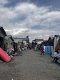 A marketplace setting with various stalls lined up on both sides of a gravel pathway. Several people are walking and browsing goods laid out at the stalls. Rolled up mats, fabrics, and other merchandise are visible. The sky is partly cloudy, adding a diffused light to the scene.