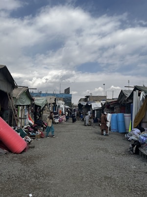 A marketplace setting with various stalls lined up on both sides of a gravel pathway. Several people are walking and browsing goods laid out at the stalls. Rolled up mats, fabrics, and other merchandise are visible. The sky is partly cloudy, adding a diffused light to the scene.