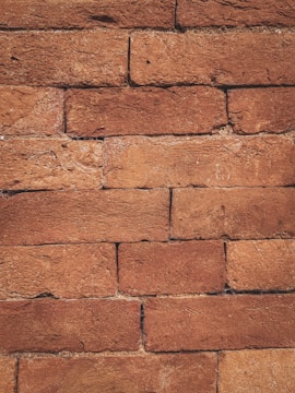 A close-up view of a brick wall constructed from rectangular red bricks. The surface of the bricks shows natural textures and slight variations in color, varying from light to dark red. The mortar between the bricks is mostly dark and blends well with the overall reddish tone of the wall.