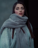 A woman is wrapped in a large, fluffy, light gray coat with a high collar. Her straight, dark hair peeks out from underneath. She has expressive eyes, and her makeup is polished, with emphasis on her eyebrows and lips. The background is blurred and dark, adding a sense of depth.