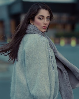 A woman with long dark hair wearing a stylish gray coat is turned slightly to the side, gazing directly at the camera. Her makeup accentuates her eyes and she appears confident and composed. The background is softly blurred, indicating an outdoor setting.