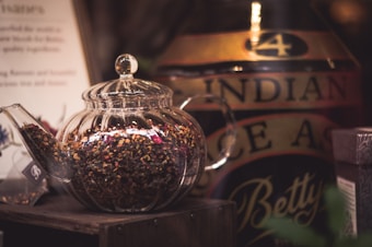 A clear glass teapot filled with loose leaf tea sits on a wooden surface. Nearby, there is a large container with the words 'Indian Black Tea' partially visible. The setting appears cozy and inviting, likely in a tea shop or kitchen.