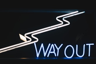 Neon sign that reads 'WAY OUT' with a zigzag arrow above it pointing to the left. The sign is illuminated in dark surroundings, highlighting the text and arrow prominently.