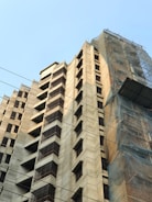 A high-rise building with a modern architectural design, featuring multiple balconies and windows. Part of the structure is covered with scaffolding and netting, indicating ongoing construction or maintenance work. Overhead cables can be seen against a clear blue sky.