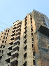 A high-rise building with a modern architectural design, featuring multiple balconies and windows. Part of the structure is covered with scaffolding and netting, indicating ongoing construction or maintenance work. Overhead cables can be seen against a clear blue sky.
