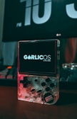 A clear handheld gaming device is placed on a desk with its screen displaying the text 'GarlicOS by Black Seraph'. The device features a D-pad, several buttons, and a transparent casing that reveals its internal components. In the background, a large digital clock displays the time as 10 PM, and part of a keyboard is visible.
