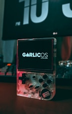 A clear handheld gaming device is placed on a desk with its screen displaying the text 'GarlicOS by Black Seraph'. The device features a D-pad, several buttons, and a transparent casing that reveals its internal components. In the background, a large digital clock displays the time as 10 PM, and part of a keyboard is visible.