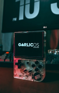 A clear handheld gaming device is placed on a desk with its screen displaying the text 'GarlicOS by Black Seraph'. The device features a D-pad, several buttons, and a transparent casing that reveals its internal components. In the background, a large digital clock displays the time as 10 PM, and part of a keyboard is visible.
