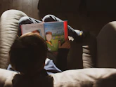 Imagem de um criança. Ela está deitada de forma bem confortável e lendo um livro infantil.