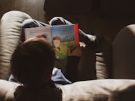 A child is sitting on a couch, engrossed in reading a colorful children's book. The child is wearing casual clothing and socks with a playful design. The room is softly lit with natural light creating a warm, cozy atmosphere.