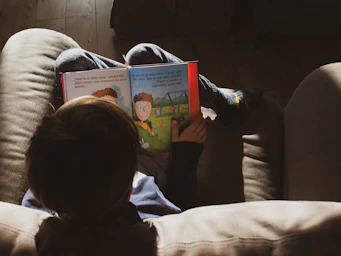 Imagem de um criança. Ela está deitada de forma bem confortável e lendo um livro infantil.