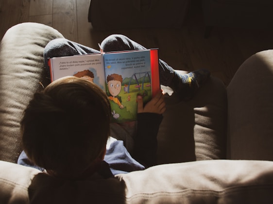 A child is sitting on a couch, engrossed in reading a colorful children's book. The child is wearing casual clothing and socks with a playful design. The room is softly lit with natural light creating a warm, cozy atmosphere.