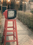 A vintage red television sits atop a tall red wooden stool in an outdoor setting, with a path leading through a garden and a building entrance in the background. The television screen displays text in a script language.