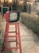 A vintage red television sits atop a tall red wooden stool in an outdoor setting, with a path leading through a garden and a building entrance in the background. The television screen displays text in a script language.