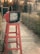 A vintage red television sits atop a tall red wooden stool in an outdoor setting, with a path leading through a garden and a building entrance in the background. The television screen displays text in a script language.