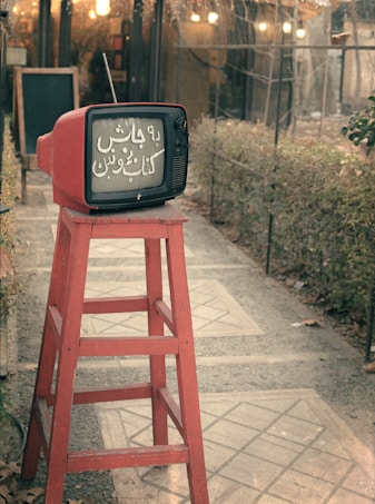 A vintage red television sits atop a tall red wooden stool in an outdoor setting, with a path leading through a garden and a building entrance in the background. The television screen displays text in a script language.